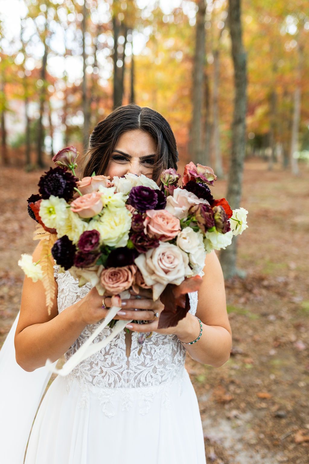 Smiling bride hiding behind colorful fall wedding bouquet of roses in forest.