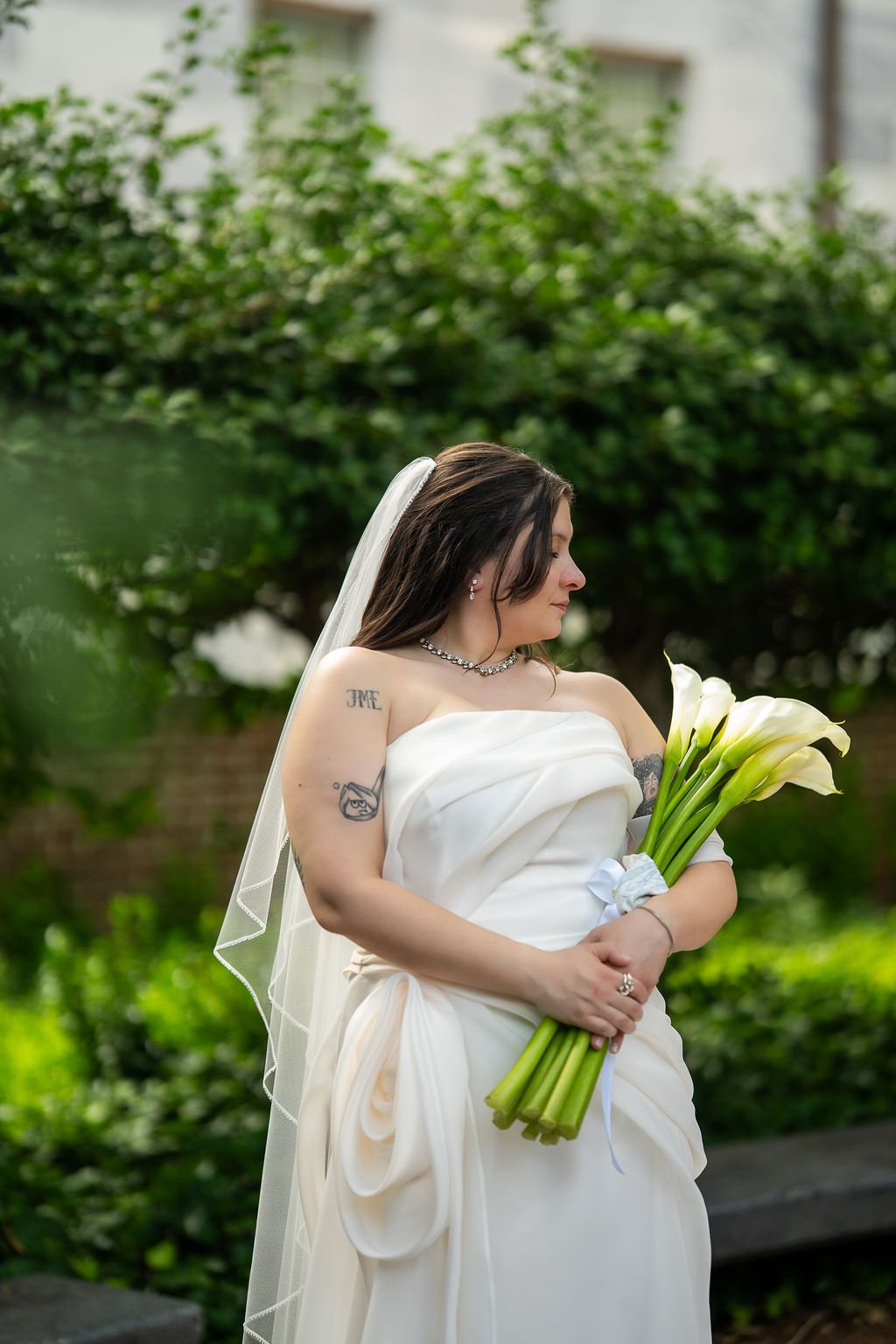 Bride in strapless white gown with veil holds calla lily bouquet while gazing down in a lush garden.