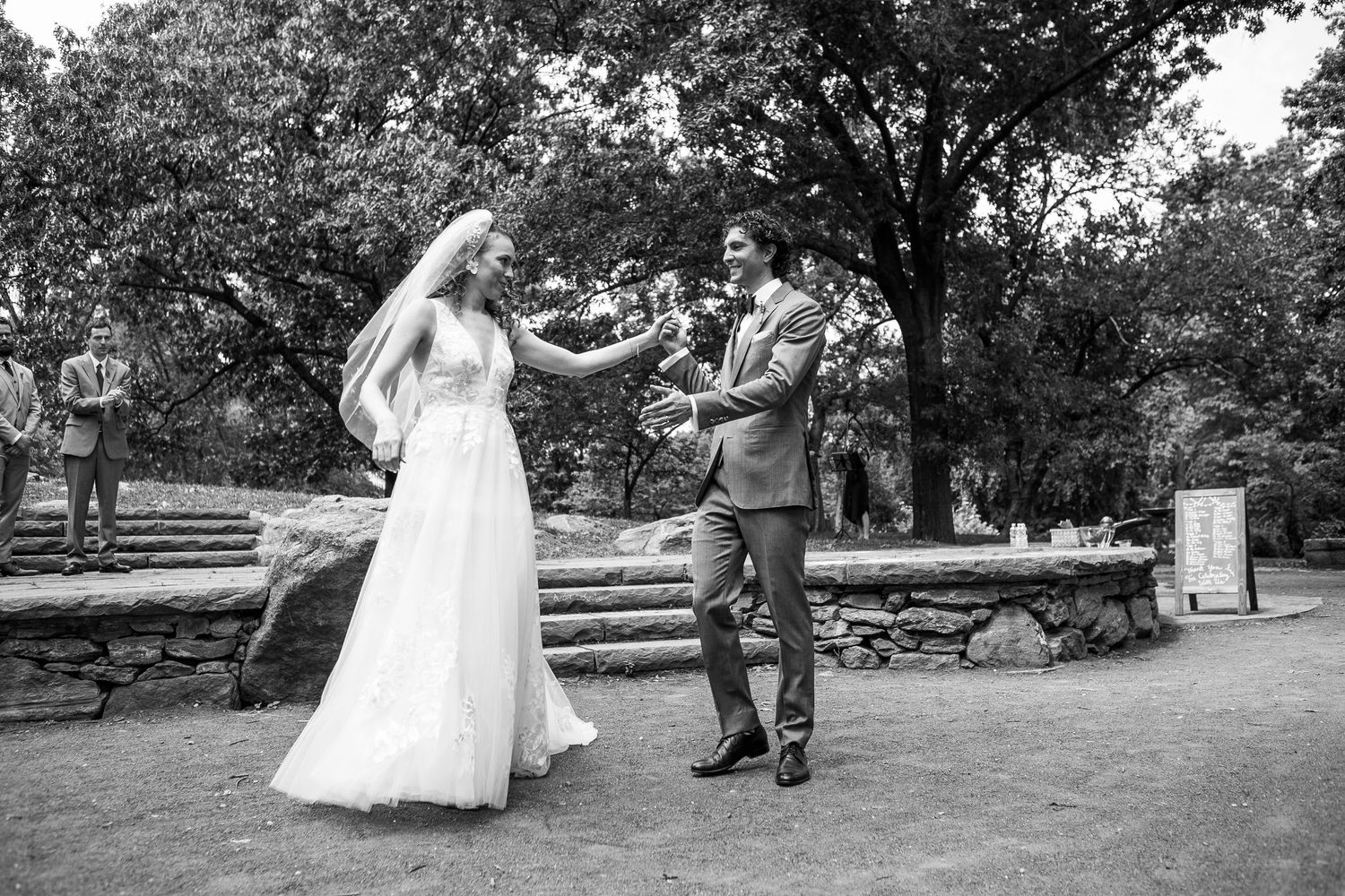 Black and white photo of bride in white gown and groom in gray suit sharing a joyful first dance outdoors.