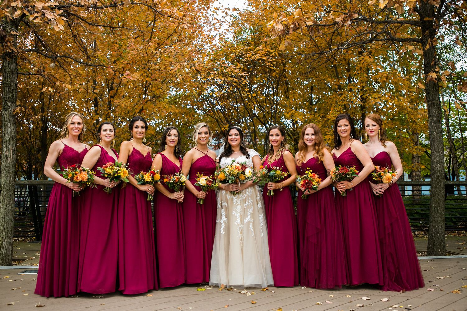Bride stands with bridesmaids in burgundy gowns holding colorful bouquets before vibrant autumn trees.