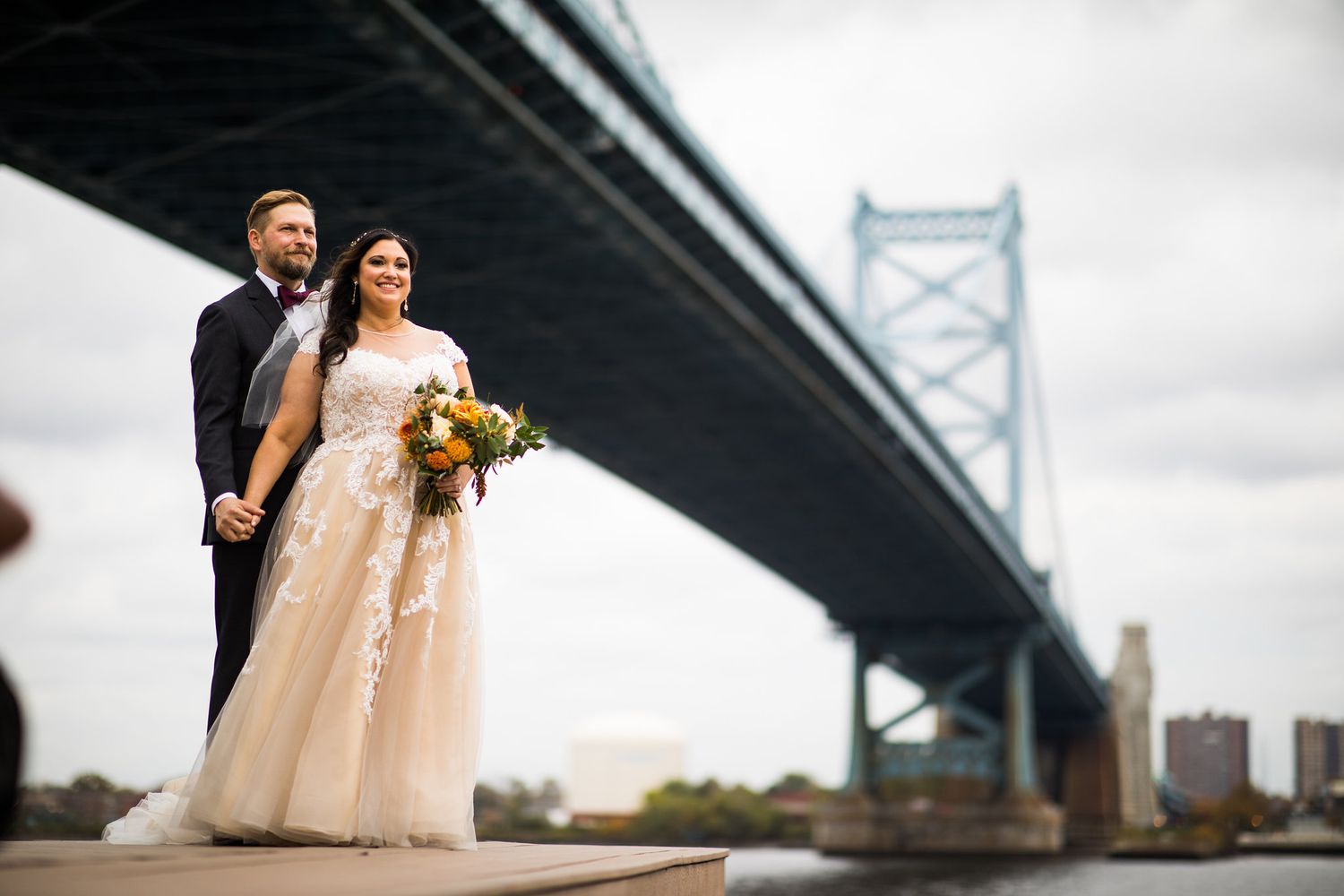 Bride and groom posing on waterfront dock with the Benjamin Franklin Bridge and cityscape behind them.