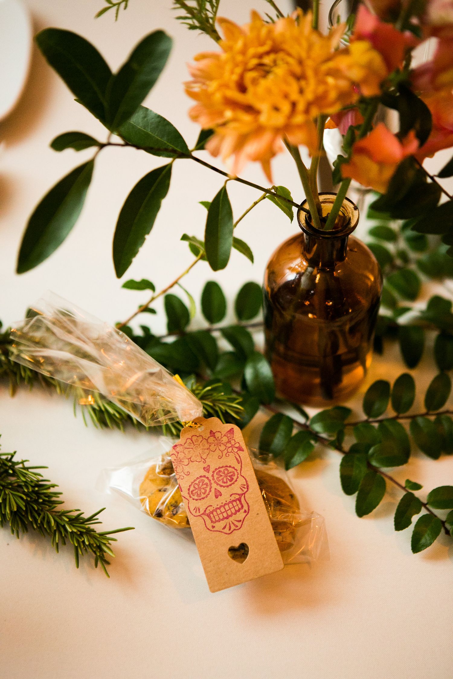 Wedding table centerpiece with orange chrysanthemum in amber bottle, greenery, and sugar skull tagged cookie favor.