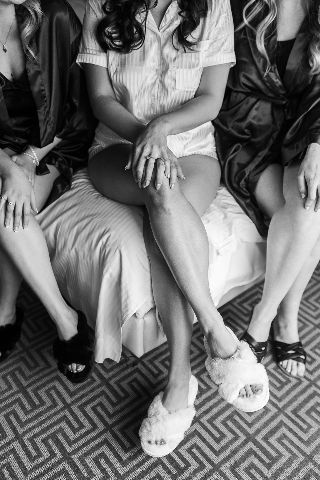 Black and white close-up of bride and bridesmaids seated on a bed with the bride wearing fluffy white slippers.
