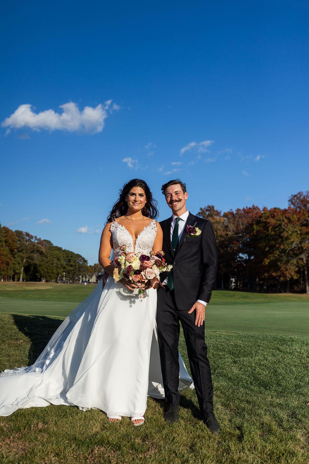 Bride in white lace gown holding bouquet stands with groom in black suit on green golf course under blue sky.