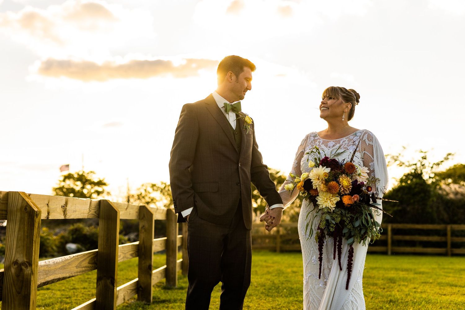 Bride and groom holding hands walking at golden hour near a wooden fence at an outdoor wedding.