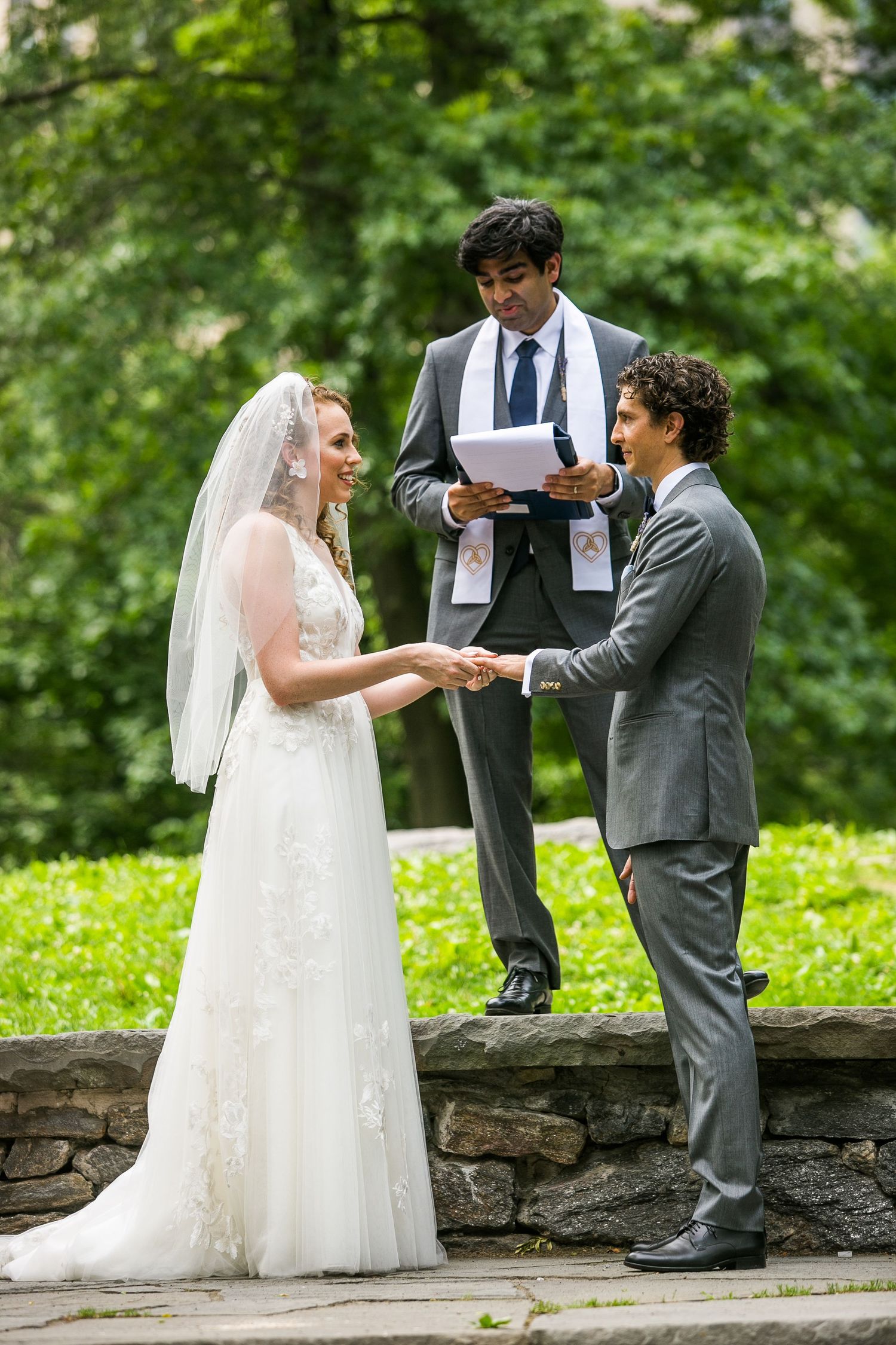 Bride and groom holding hands during outdoor wedding ceremony with officiant reading from a book.