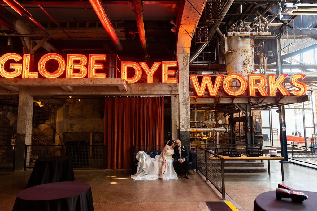 Bride and groom sit beneath glowing Globe Dye Works sign in industrial wedding venue.