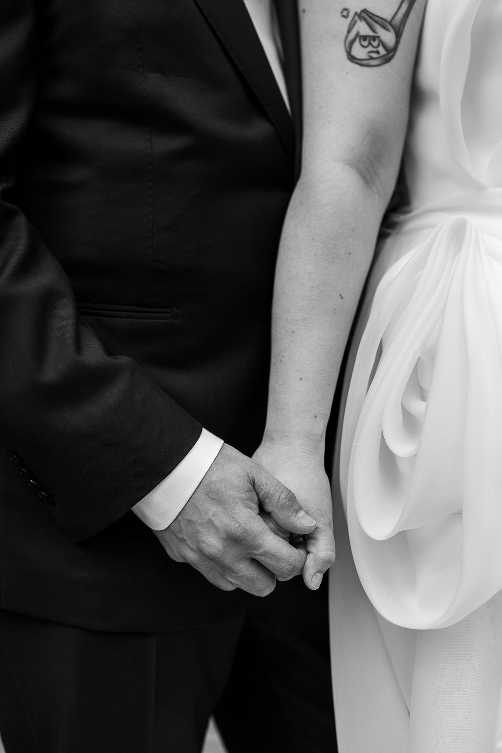 Close-up black and white photo of bride and groom holding hands, showing tattoo on brides arm and wedding attire details.