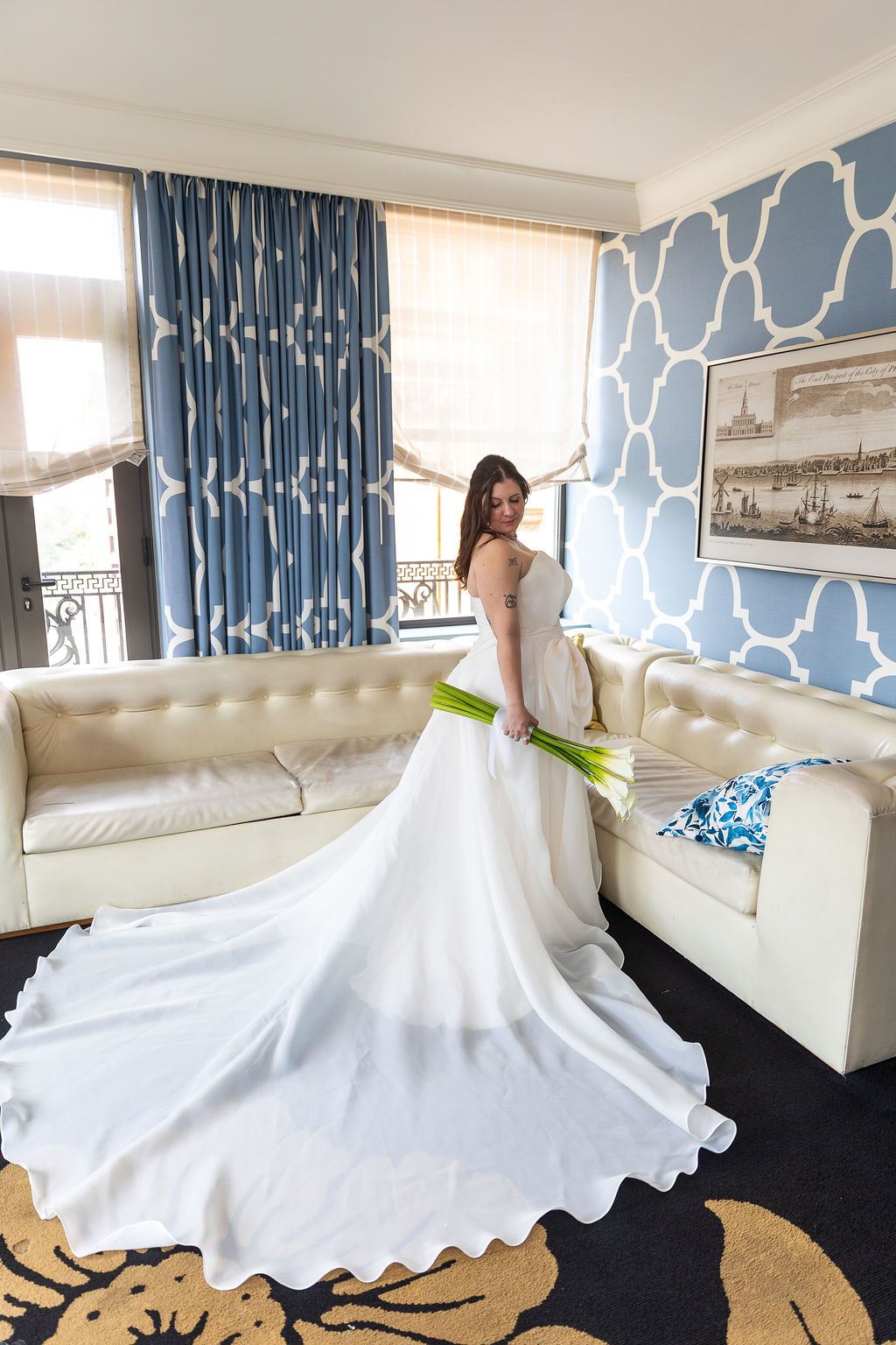 Bride in stunning white wedding gown with cathedral train holding green bouquet in a blue hotel suite.