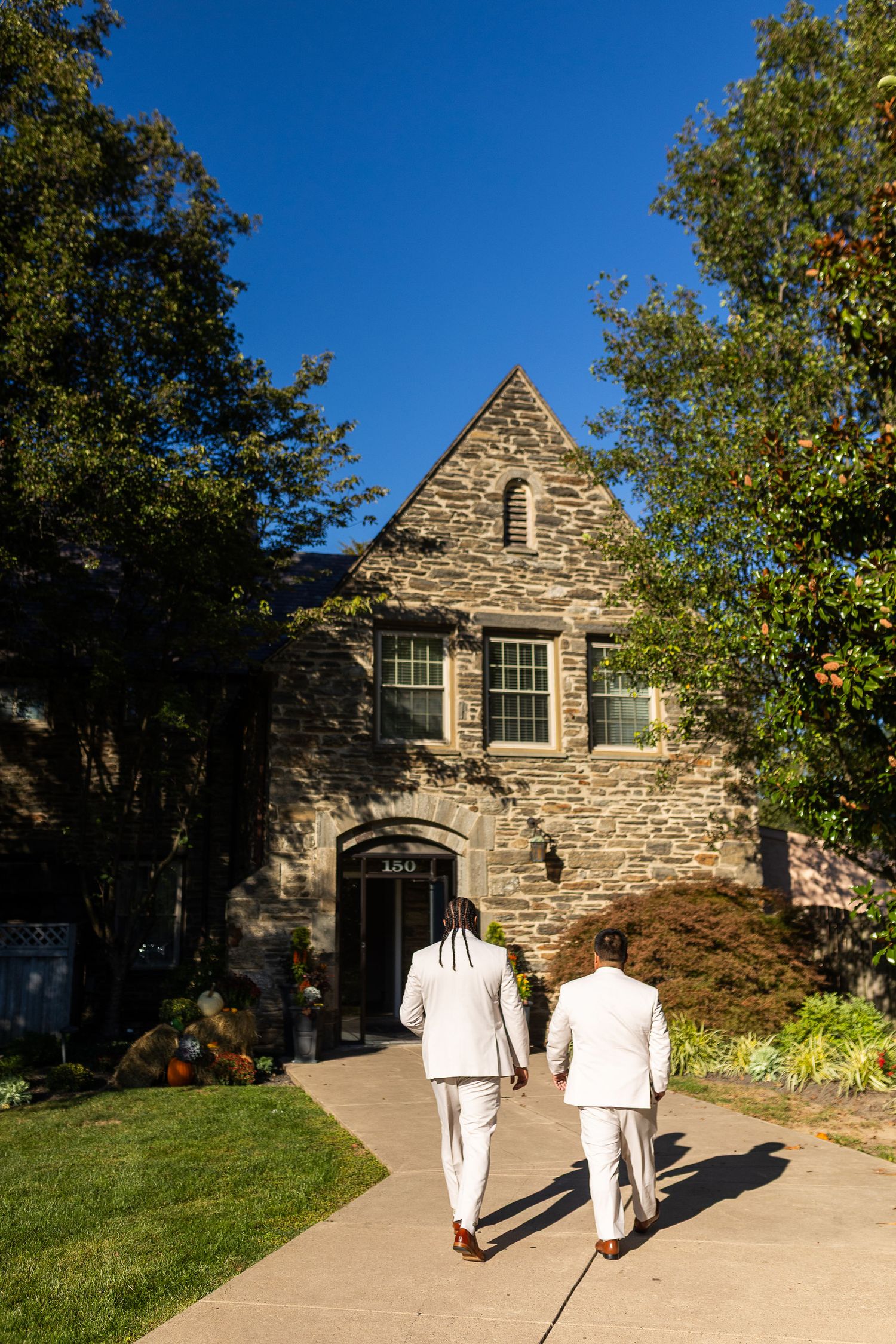 Two grooms in white suits walking toward stone building on sunny wedding day.
