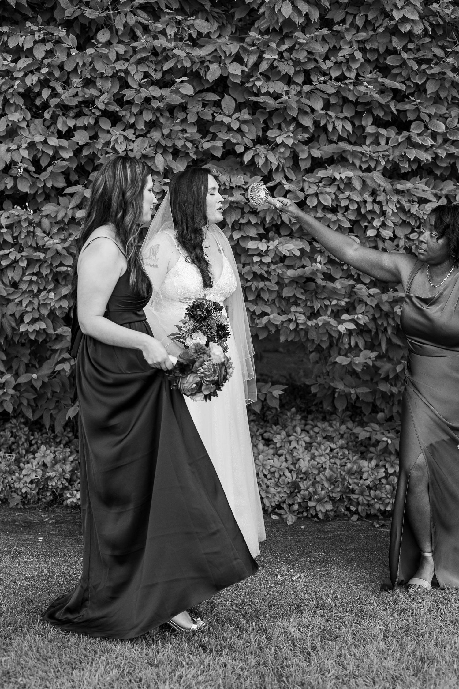 Black and white photo of a bridesmaid touching the brides face while another bridesmaid looks on outdoors.