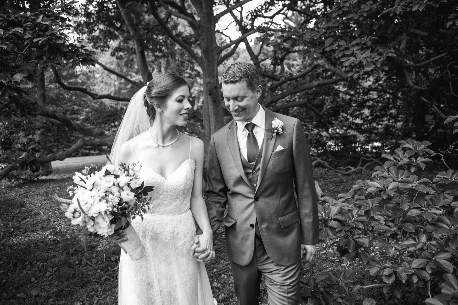 Black and white photo of bride and groom walking hand in hand through a lush garden setting.