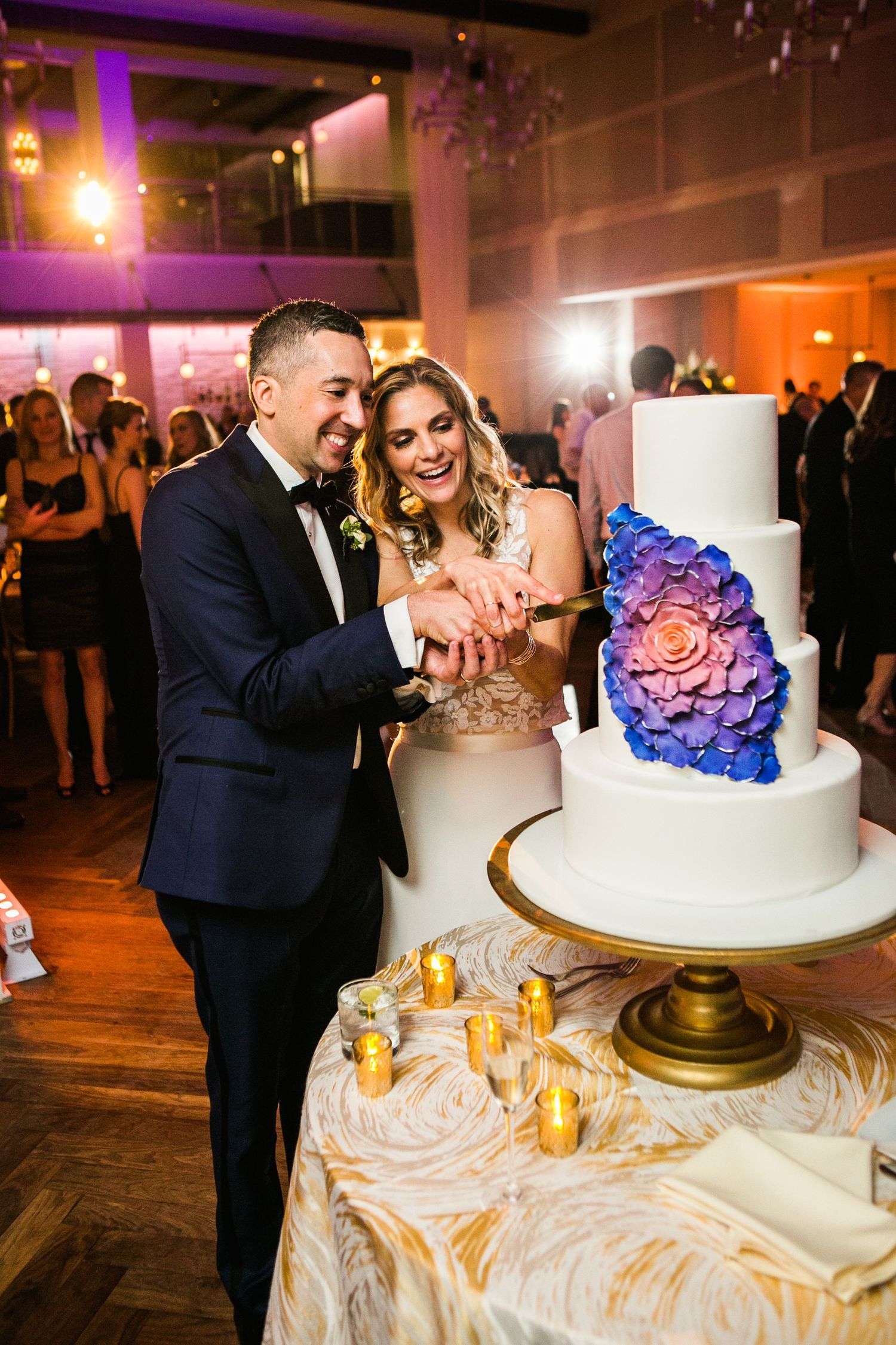 Smiling bride and groom cutting a white three-tier wedding cake with blue and purple floral details.