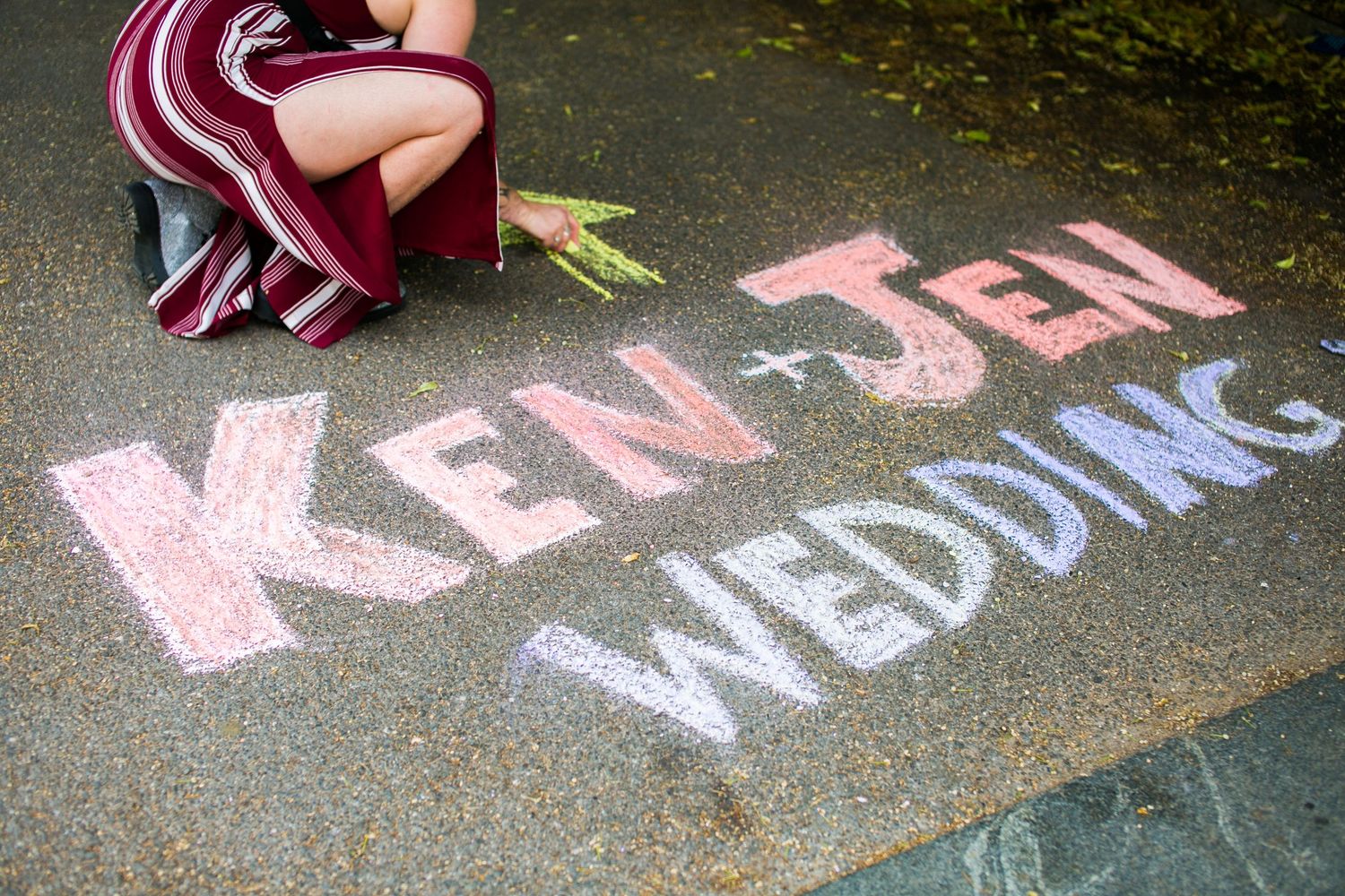 Colorful chalk writing on pavement reading Ken and Jen Wedding with a person crouching nearby.