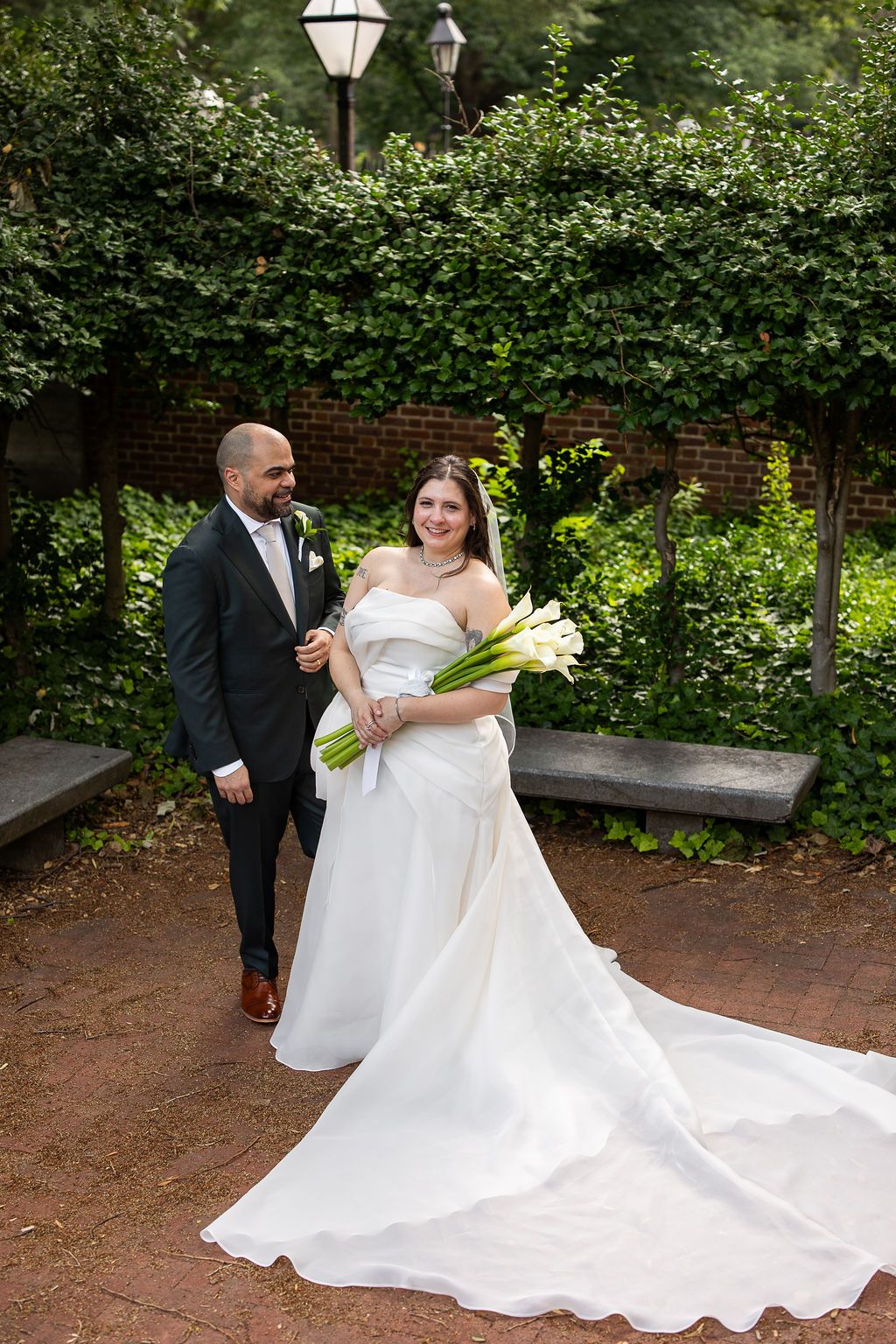 Bride holding white calla lily bouquet in elegant gown with long train walks beside groom in brick garden.