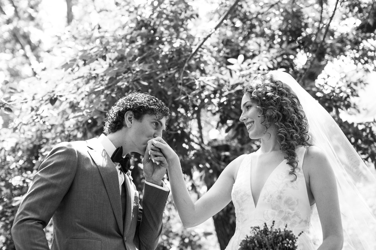 Black and white photo of groom kissing bride's hand as she smiles holding a bouquet outdoors.