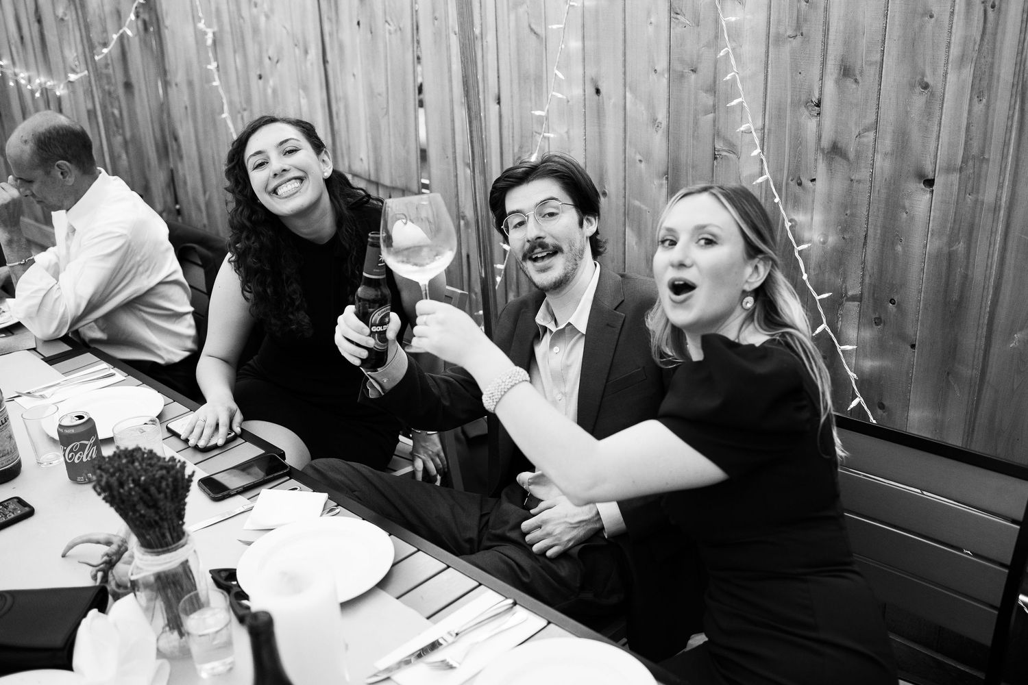 Black and white photo of three wedding guests laughing and toasting drinks at a reception dinner table.