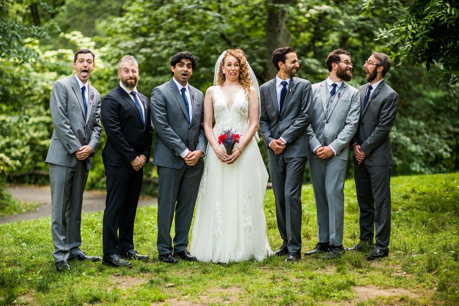 Smiling bride holding red bouquet stands with five groomsmen in matching gray suits in a green park.