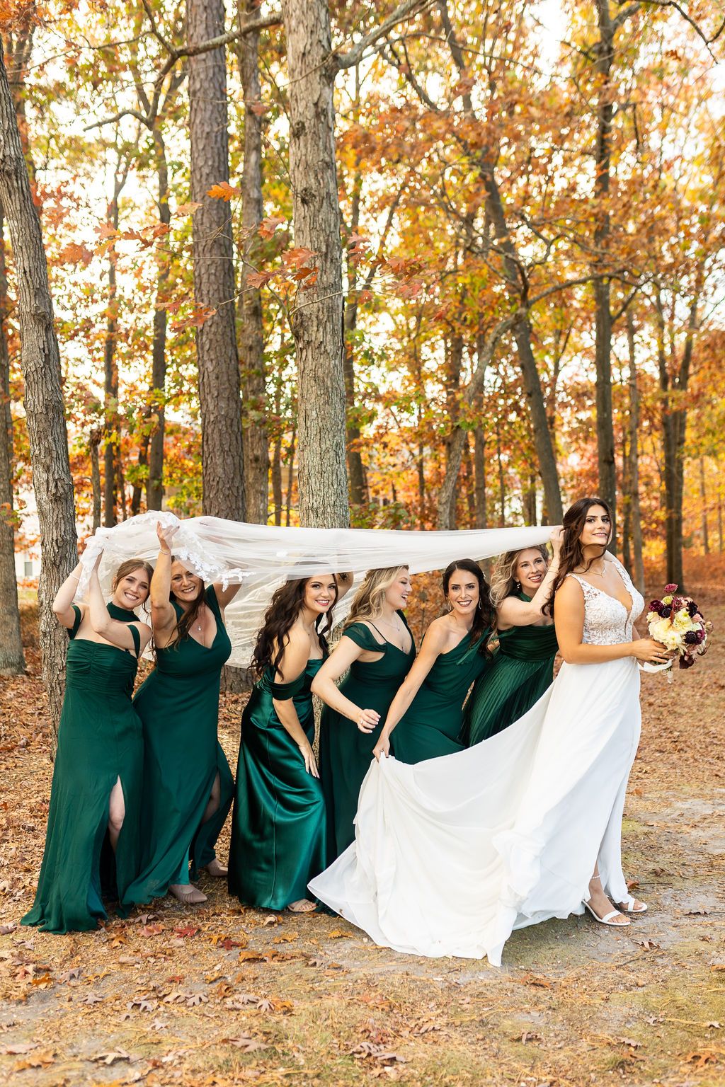 Bridesmaids in emerald green dresses shelter under bride's veil among vibrant fall foliage in the woods.