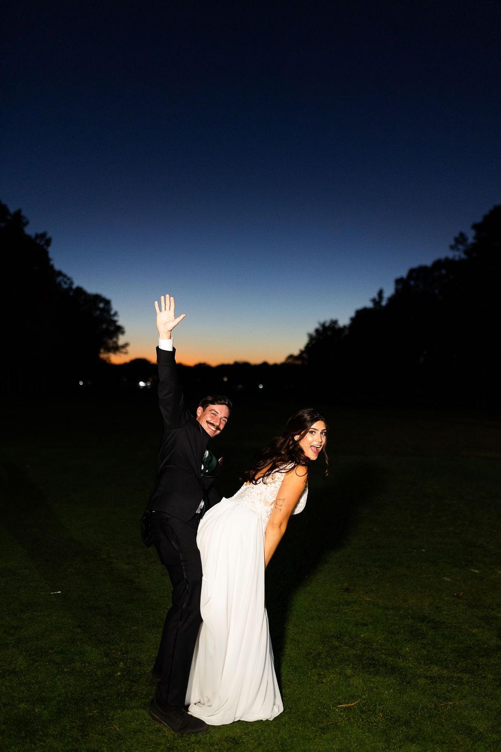Bride and groom celebrating outdoors at dusk with vibrant sunset sky behind them.