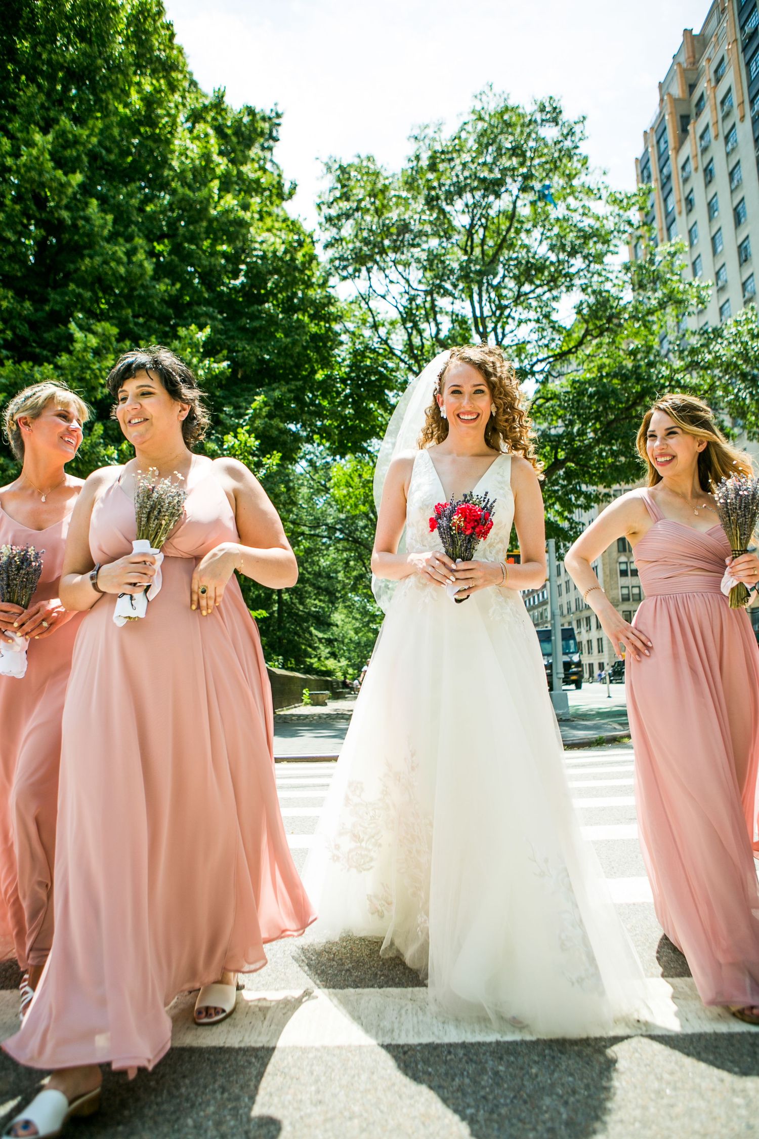 Joyful bride in white dress and veil walks with bridesmaids in pink gowns on a bright sunny day.