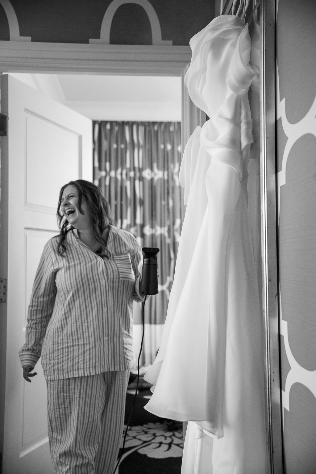 Black and white photo of bride in striped pajamas smiling while steaming her white wedding gown on a hanger.