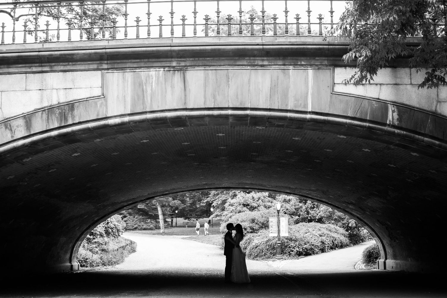 Black and white silhouette of bride and groom kissing beneath a stone bridge arch in a park.