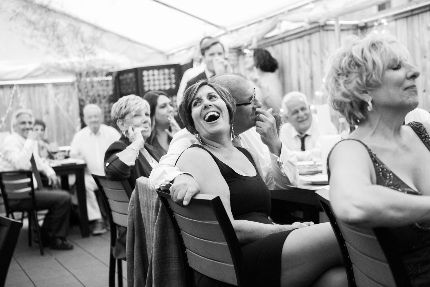Wedding guests laugh and applaud during outdoor reception under white tent in black and white photo.