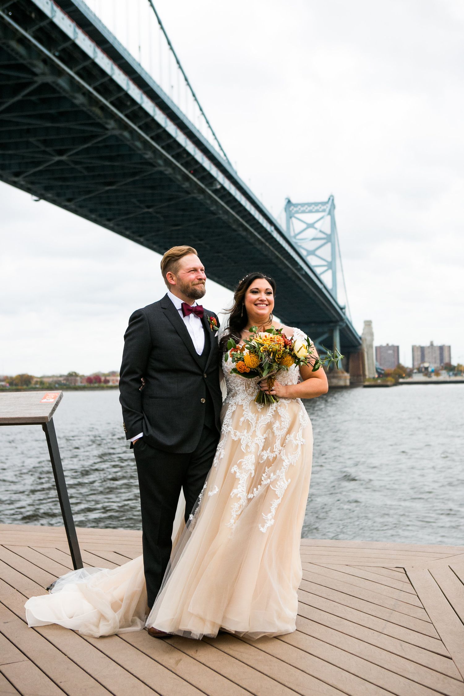 Bride holding colorful bouquet and groom in black tuxedo pose on wooden deck with Benjamin Franklin Bridge behind.