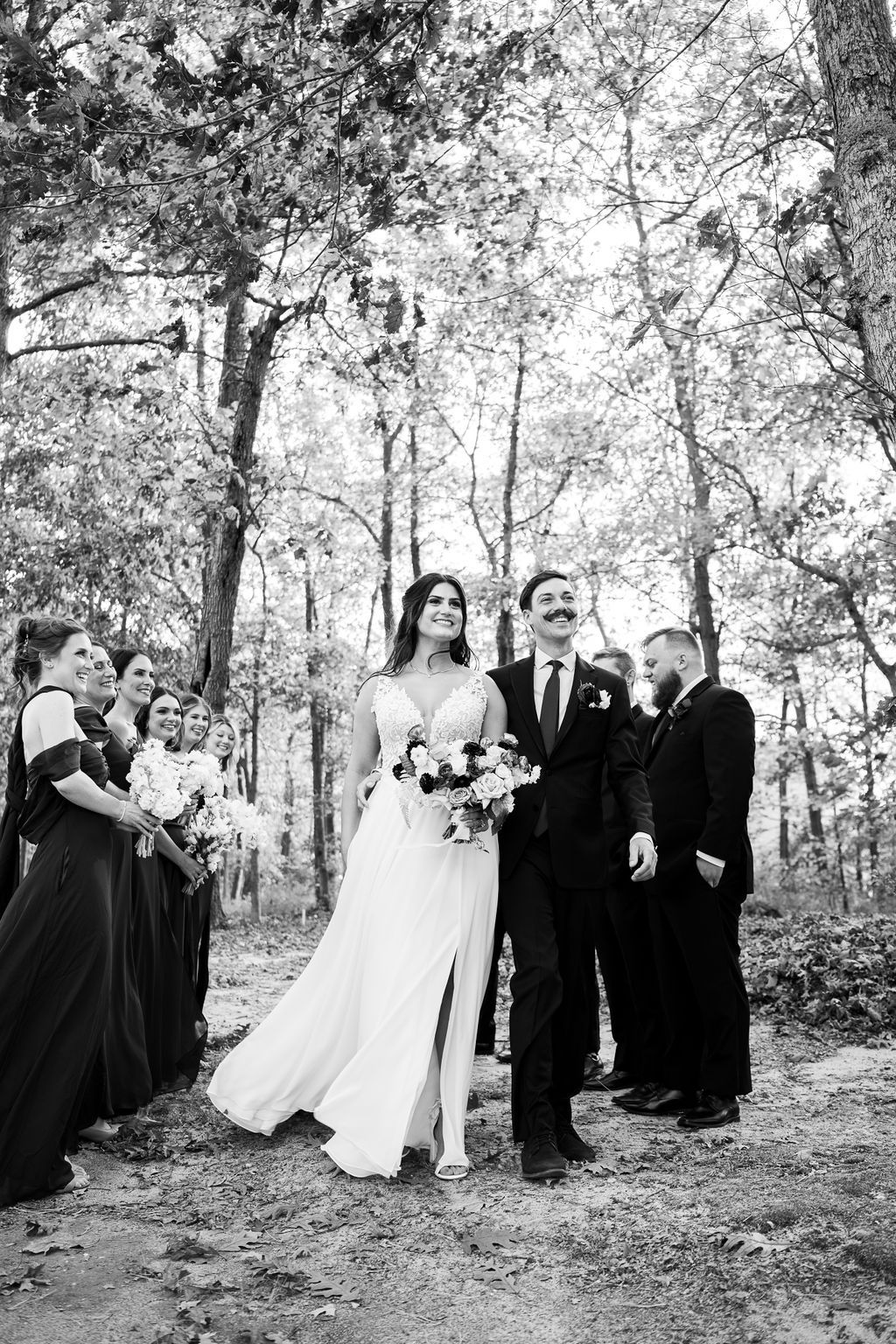 Bride and groom walking with wedding party through a wooded forest in black and white photo.