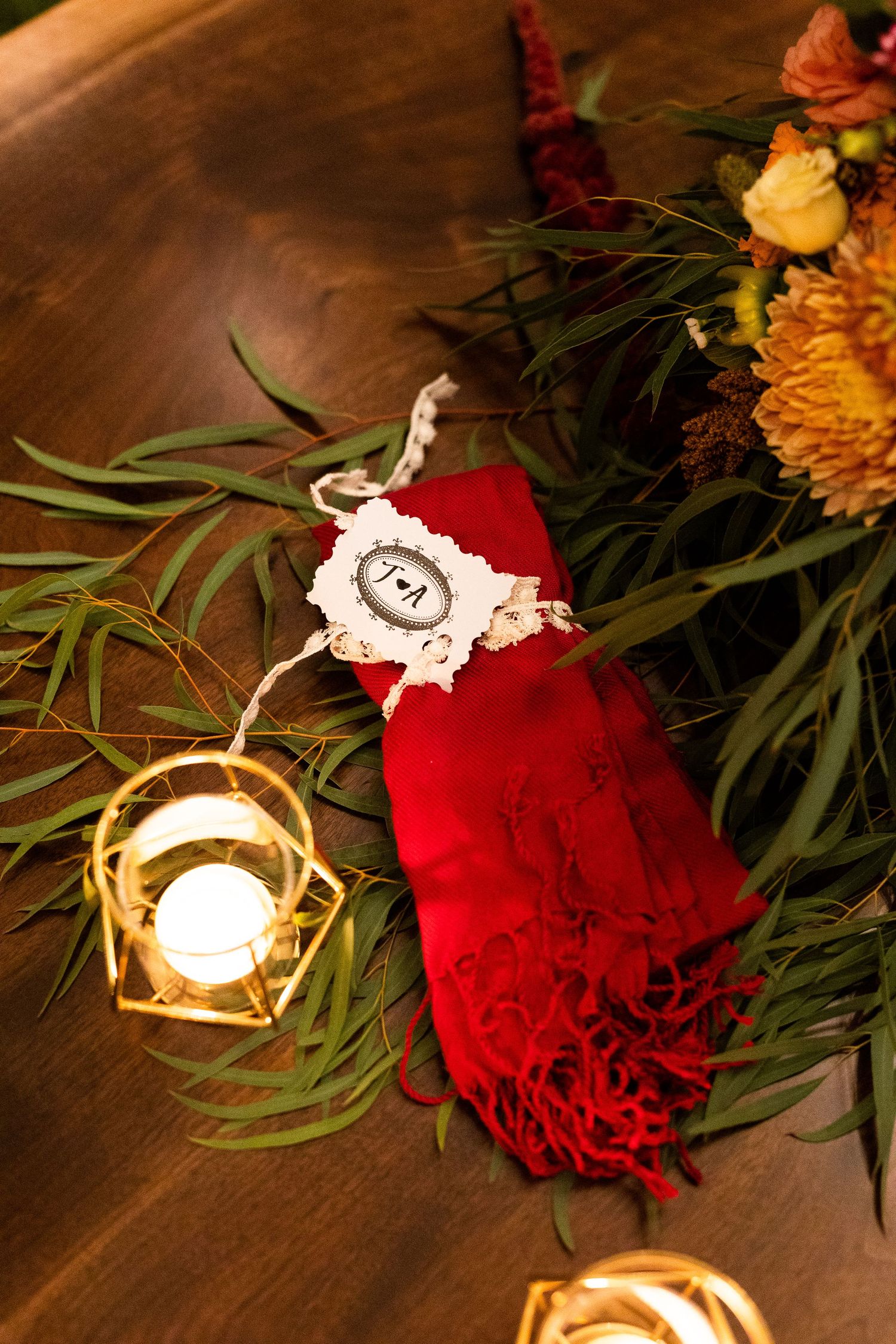 Red folded napkin with monogram tag beside gold geometric candle holder and floral greenery.