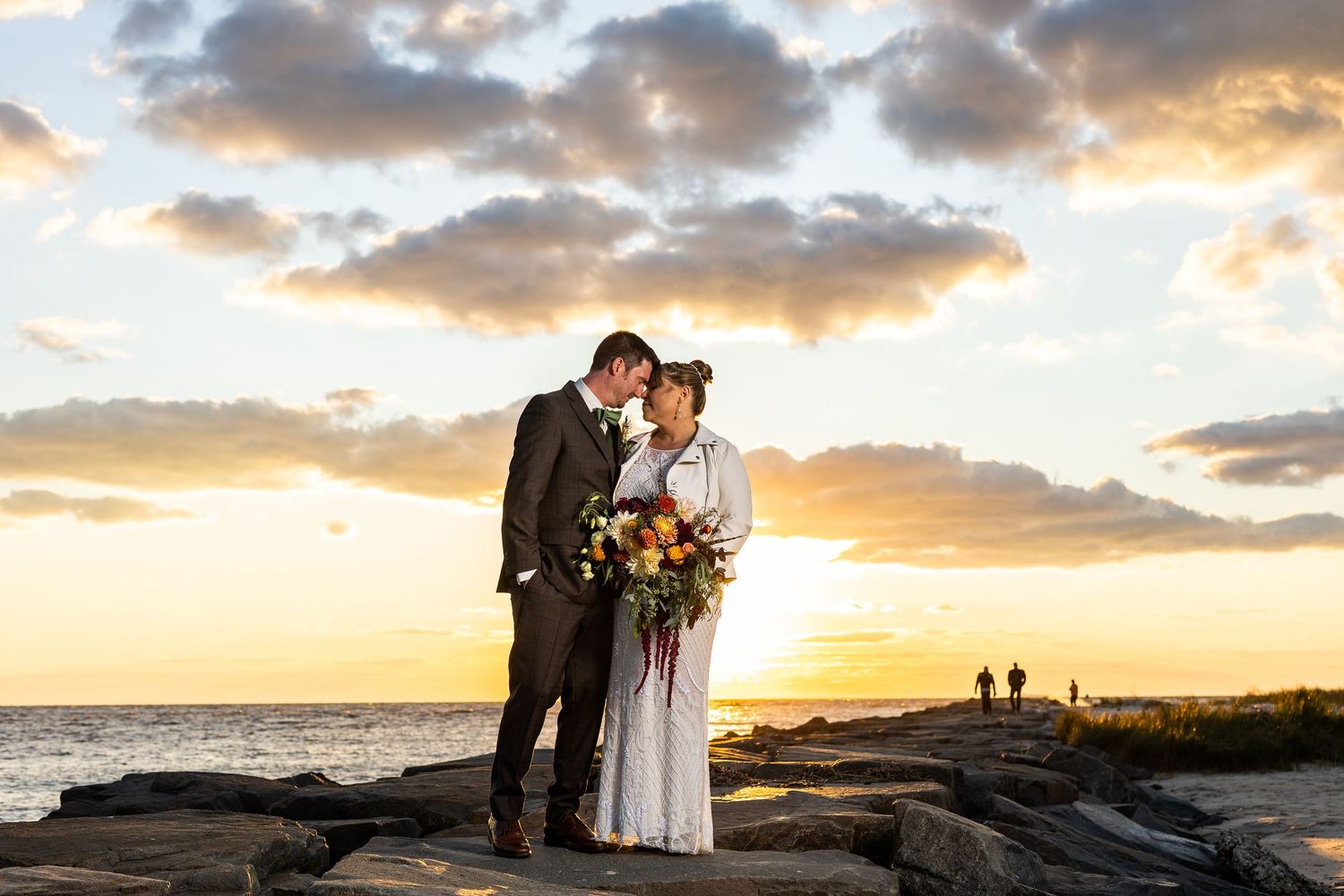 Bride and groom share a kiss on rocky shoreline with stunning golden ocean sunset backdrop.
