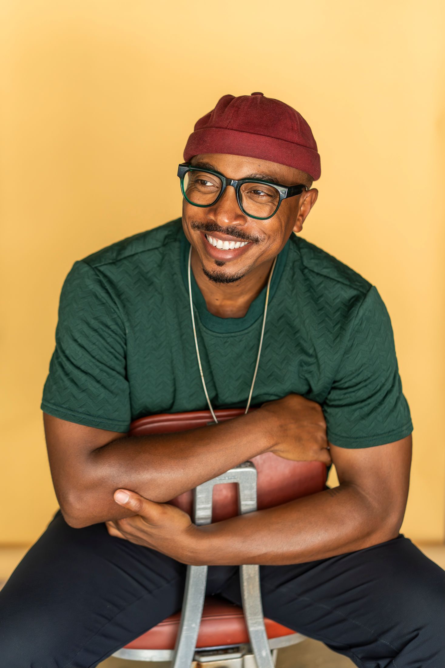 Smiling man in green shirt, red beanie, and glasses sits relaxed on a stool against a yellow background.