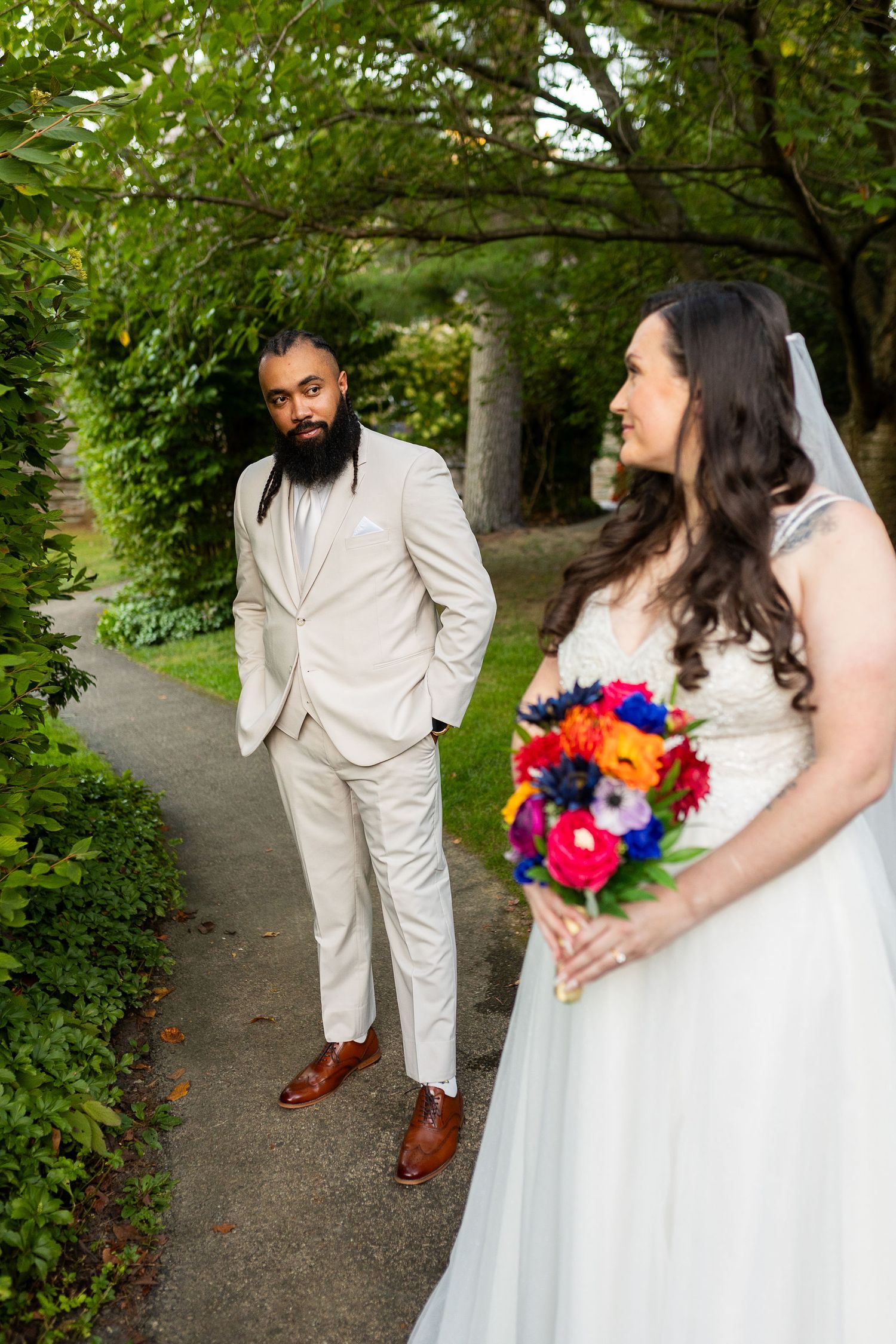 Groom in beige suit gazes at bride in white gown holding colorful bouquet on garden path.