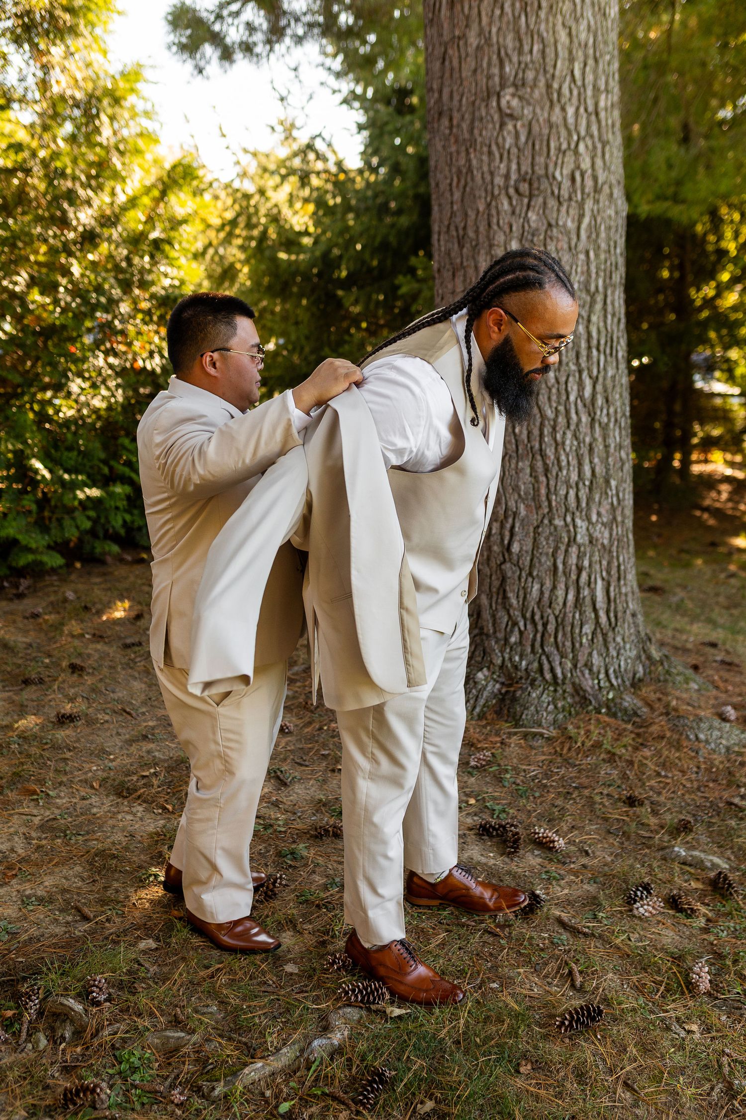 Groomsman helping bearded groom with dreadlocks put on a cream wedding suit jacket near pine trees.
