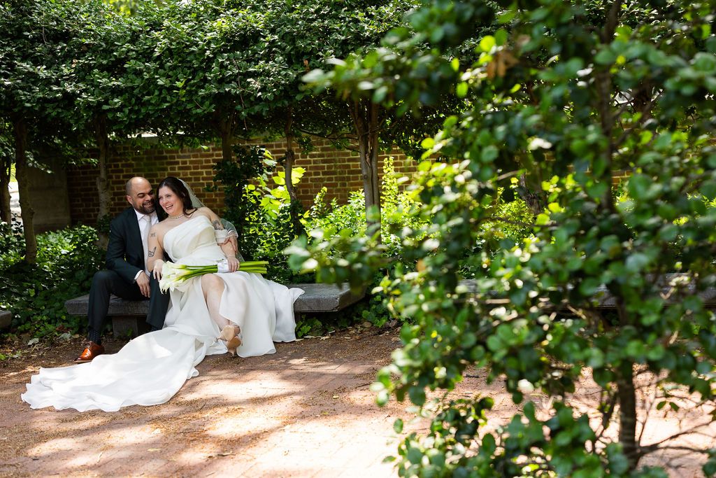 Bride and groom sitting together on a stone bench surrounded by lush green garden foliage and brick wall.
