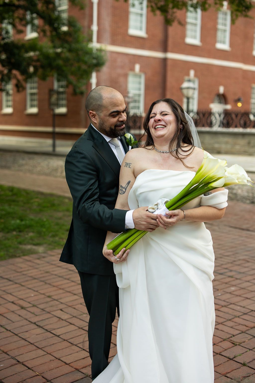 Smiling bride in white strapless gown and groom in dark suit share a joyful moment holding calla lily bouquet outdoors.