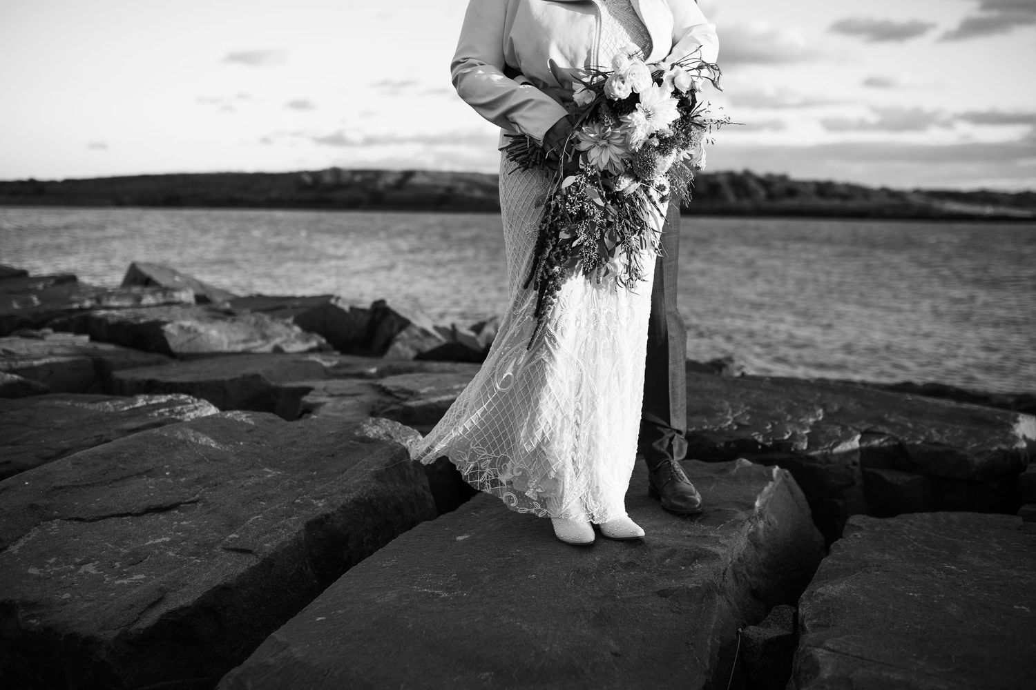 Black and white photo of bride and groom standing on rocky shore holding a large floral bouquet.