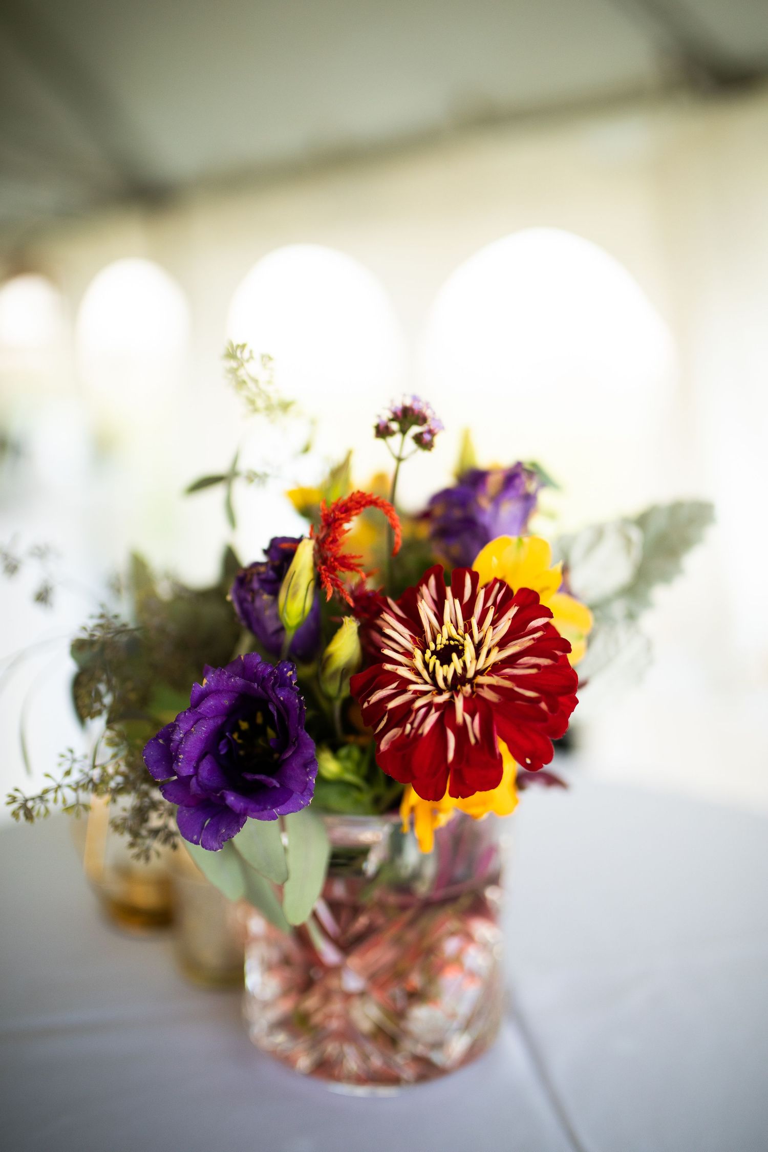 Colorful wedding centerpiece with red dahlia, purple lisianthus, and yellow flowers in a glass vase under a tent.