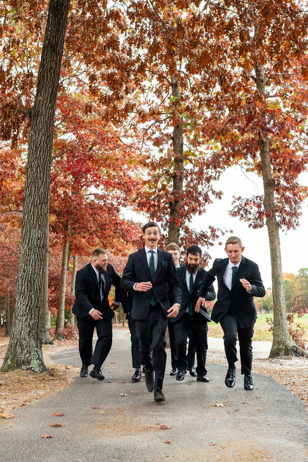 Groomsmen in black suits running playfully down a tree-lined path with vibrant autumn foliage.