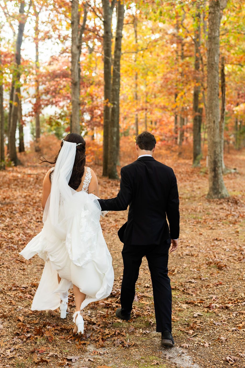 Bride and groom walk hand-in-hand through colorful autumn forest on their wedding day.