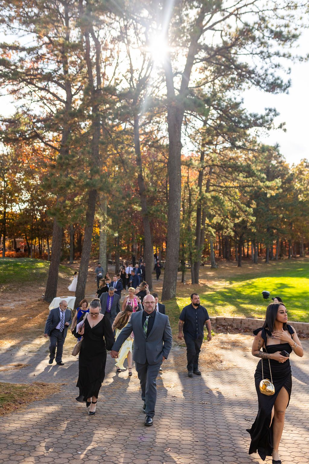 Wedding guests in formal attire walking along a sunlit brick path through tall autumn trees.