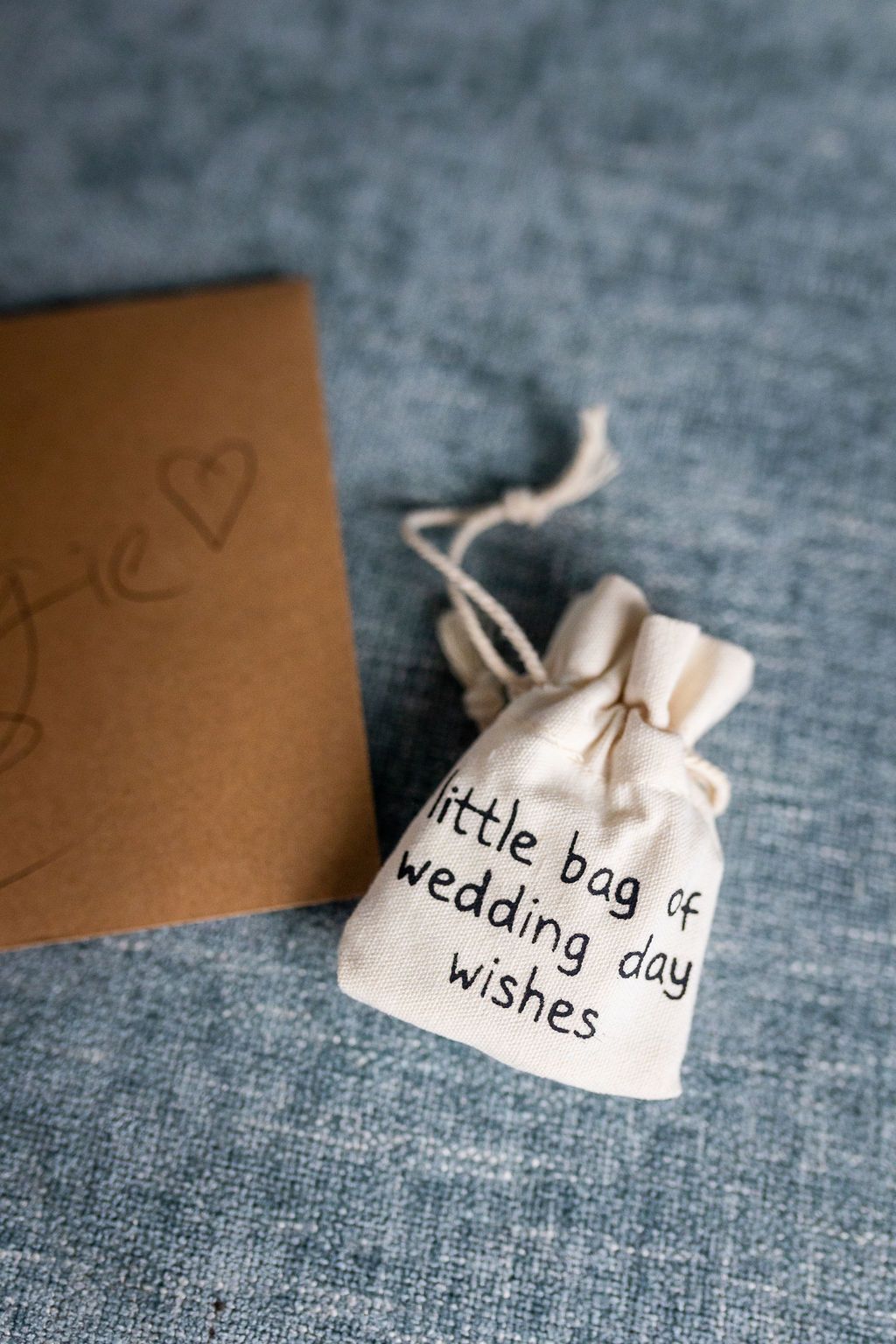 A small linen drawstring bag printed with wedding day wishes next to a kraft paper envelope on a blue surface.