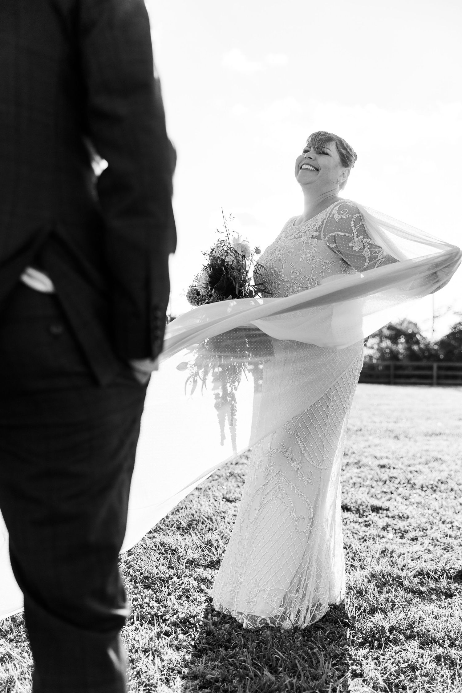 Black and white photo of laughing bride in lace gown with flowing veil billowing in an open field.