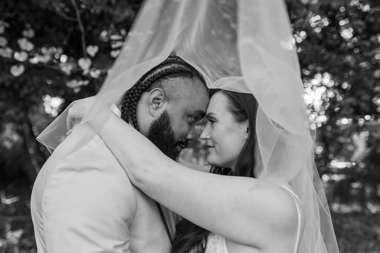 Black and white photo of bride and groom sharing intimate moment under bridal veil outdoors.
