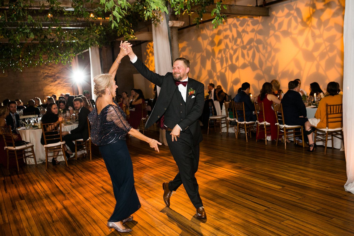A groom in a tuxedo spins his mother on the dance floor at a lively wedding reception.