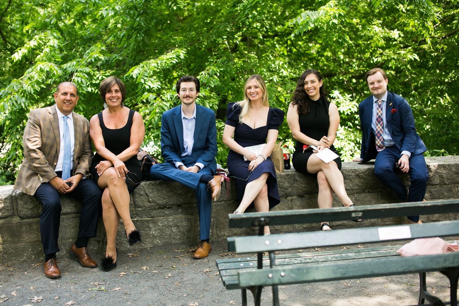 Group of six smiling wedding guests in formal wear seated on a stone wall surrounded by trees.