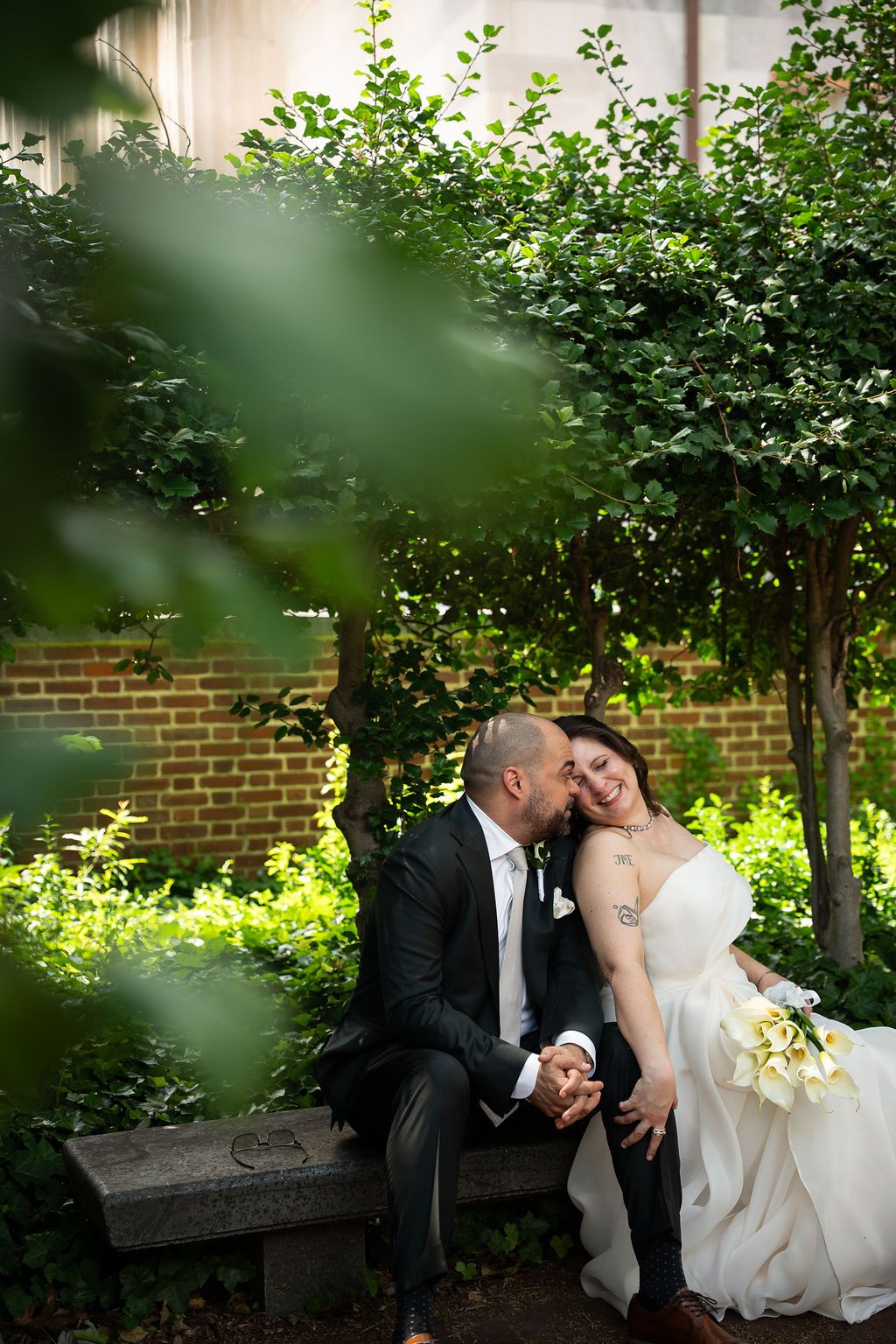 Groom kissing bride's cheek as she laughs joyfully on a bench, framed by green leaves in a garden setting.