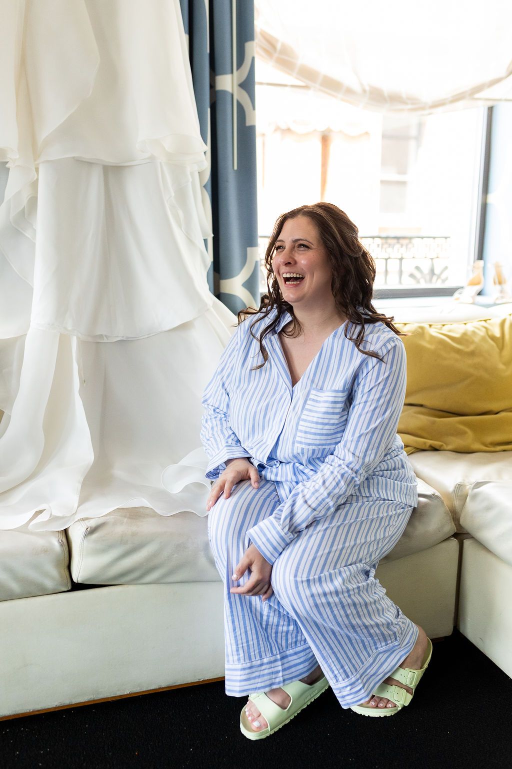 Smiling bride in blue striped pajamas and green slides sits on a cream sofa near her wedding dress.