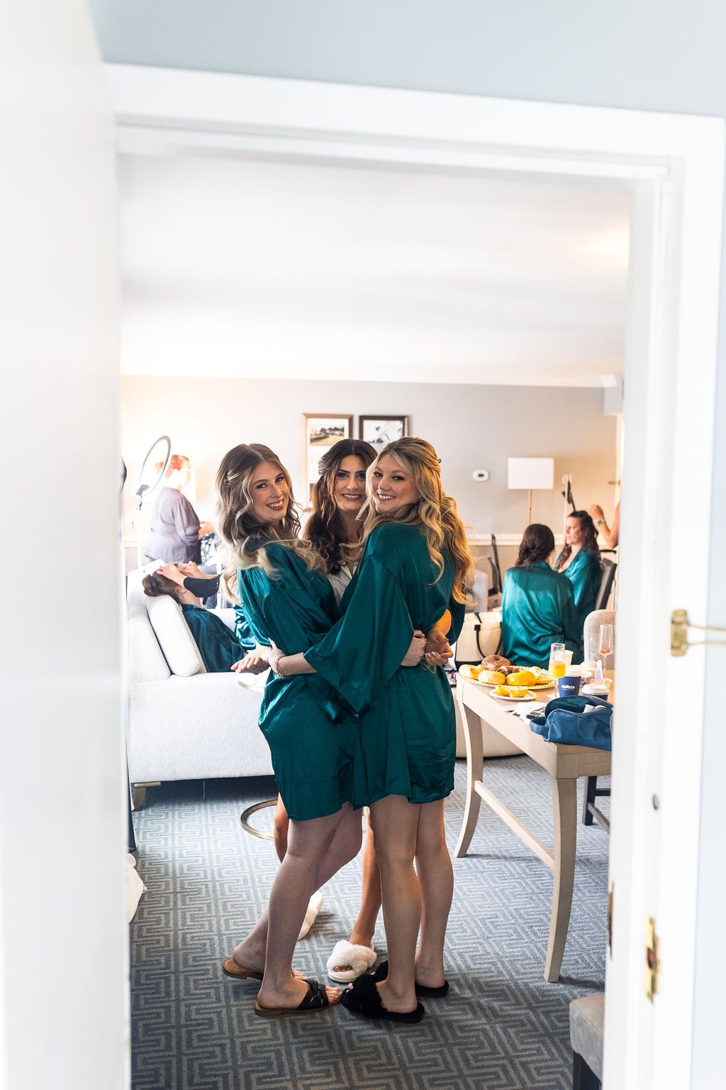 Bridesmaids in teal satin robes smiling and posing in a hotel suite while getting ready for a wedding.